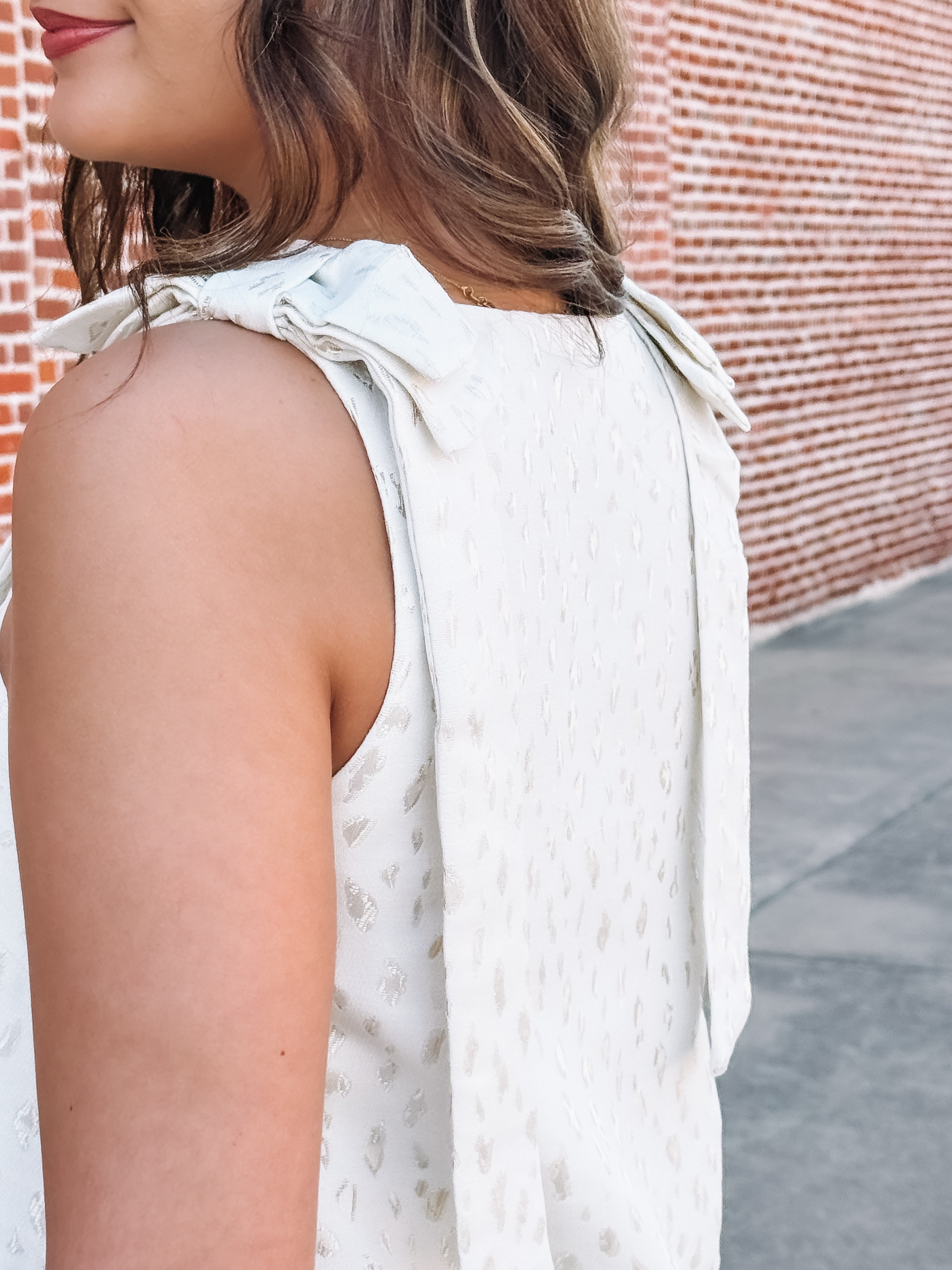 Woman wearing a white sleeveless top with bow details against a brick wall.