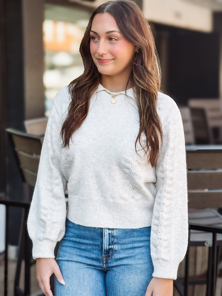 Woman wearing a cream sweater and blue jeans standing outdoors.