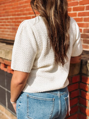 Person wearing a textured white top and blue jeans against a brick wall.