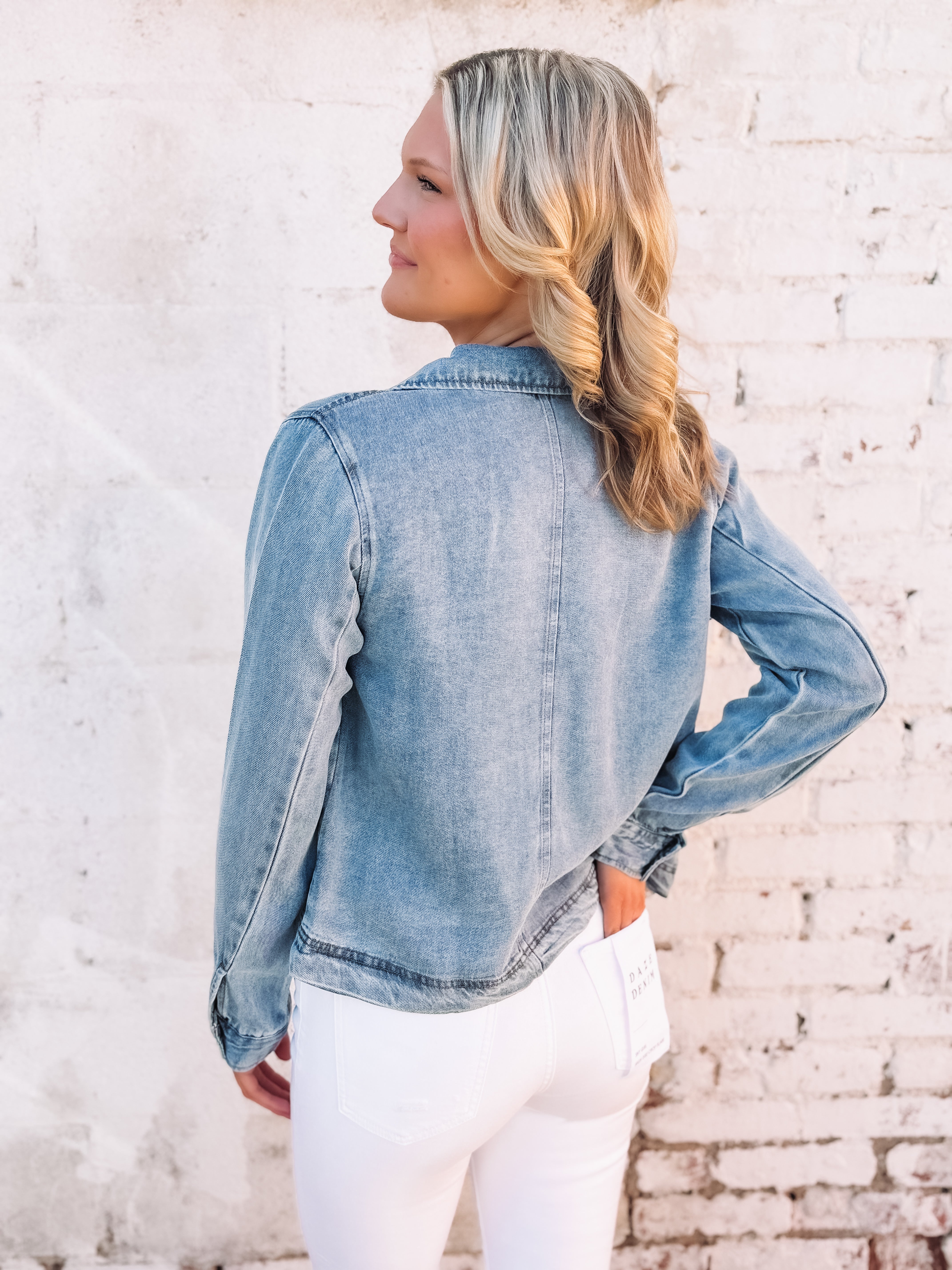 Woman wearing a soft denim hued jacket over a gray top and white pants against a light brick wall.