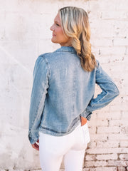 Woman wearing a soft denim hued jacket over a gray top and white pants against a light brick wall.