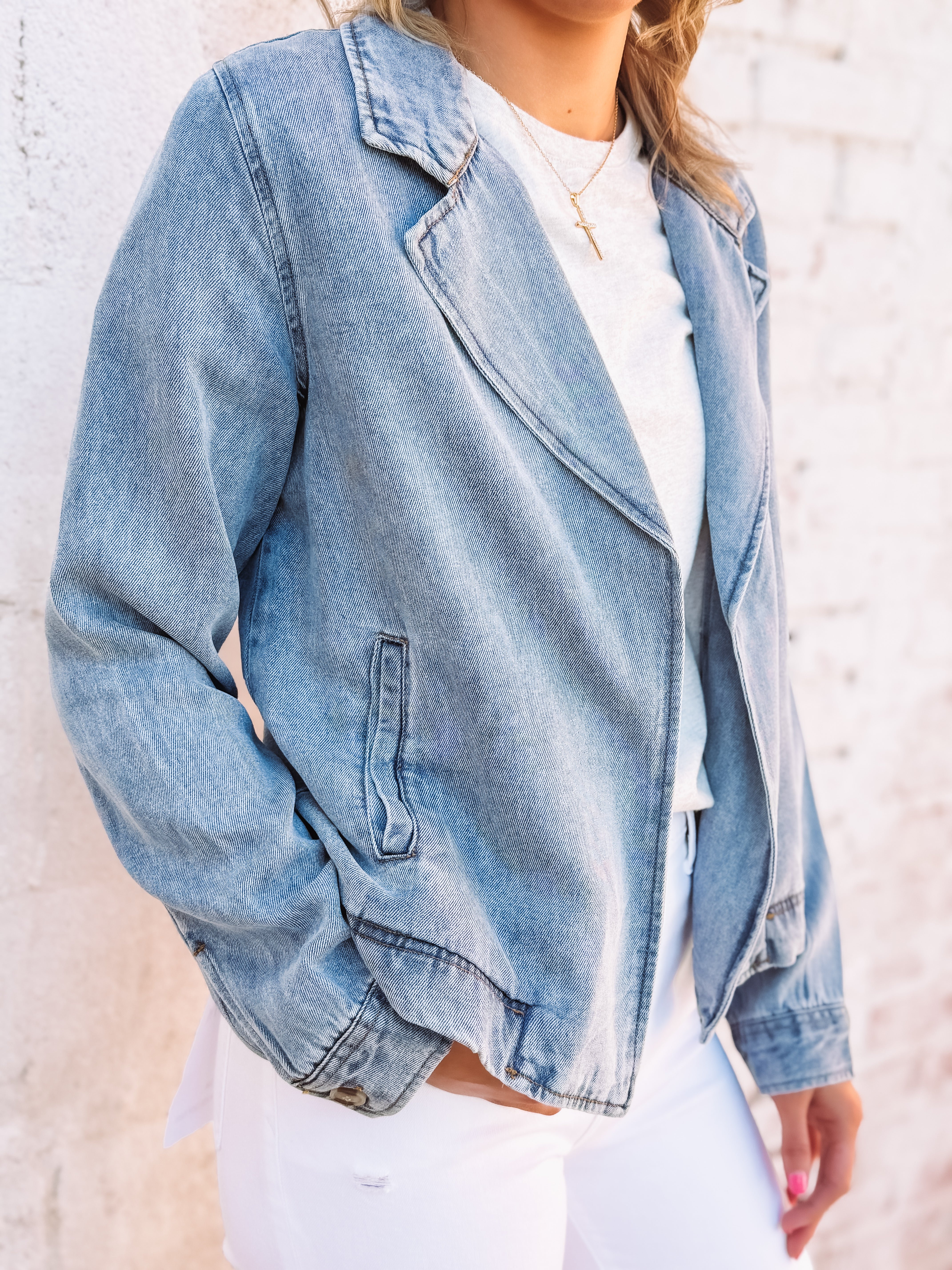 Woman wearing a soft denim hued jacket over a gray top and white pants against a light brick wall.