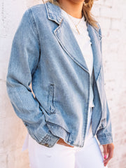 Woman wearing a soft denim hued jacket over a gray top and white pants against a light brick wall.