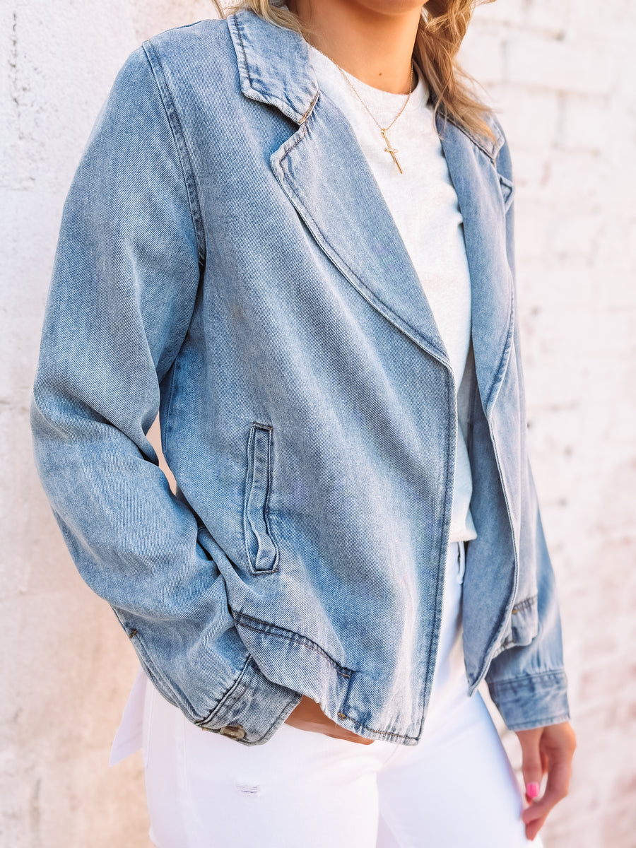 Woman wearing a soft denim hued jacket over a gray top and white pants against a light brick wall.