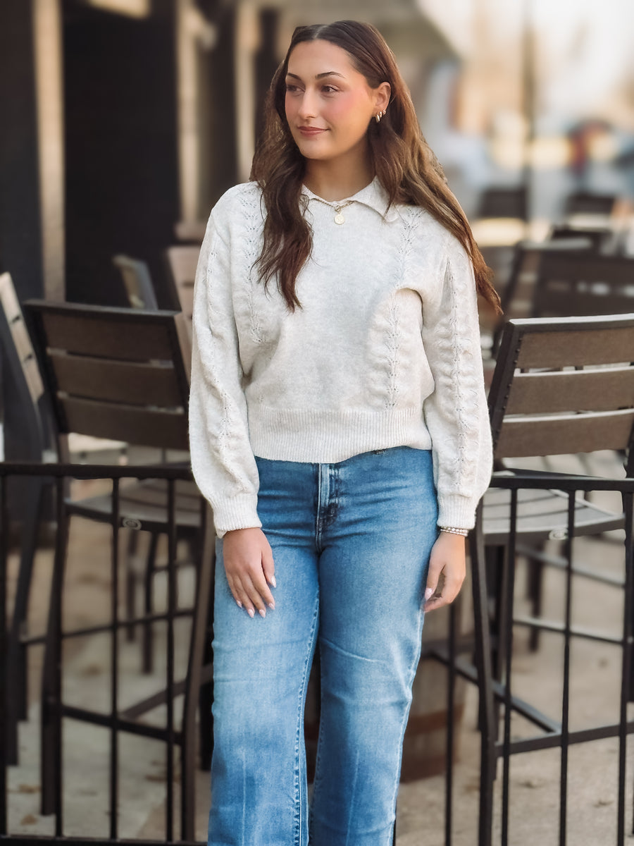 Woman wearing a cream sweater and blue jeans standing outdoors.