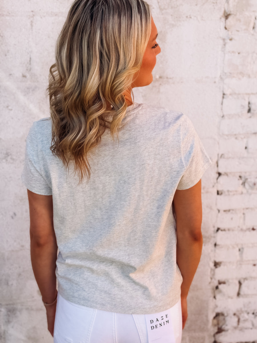 Woman wearing a light gray t-shirt with 'Daze Denim' branding against a textured white wall.