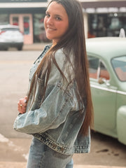 Woman wearing a denim jacket standing in front of a vintage car on a street.