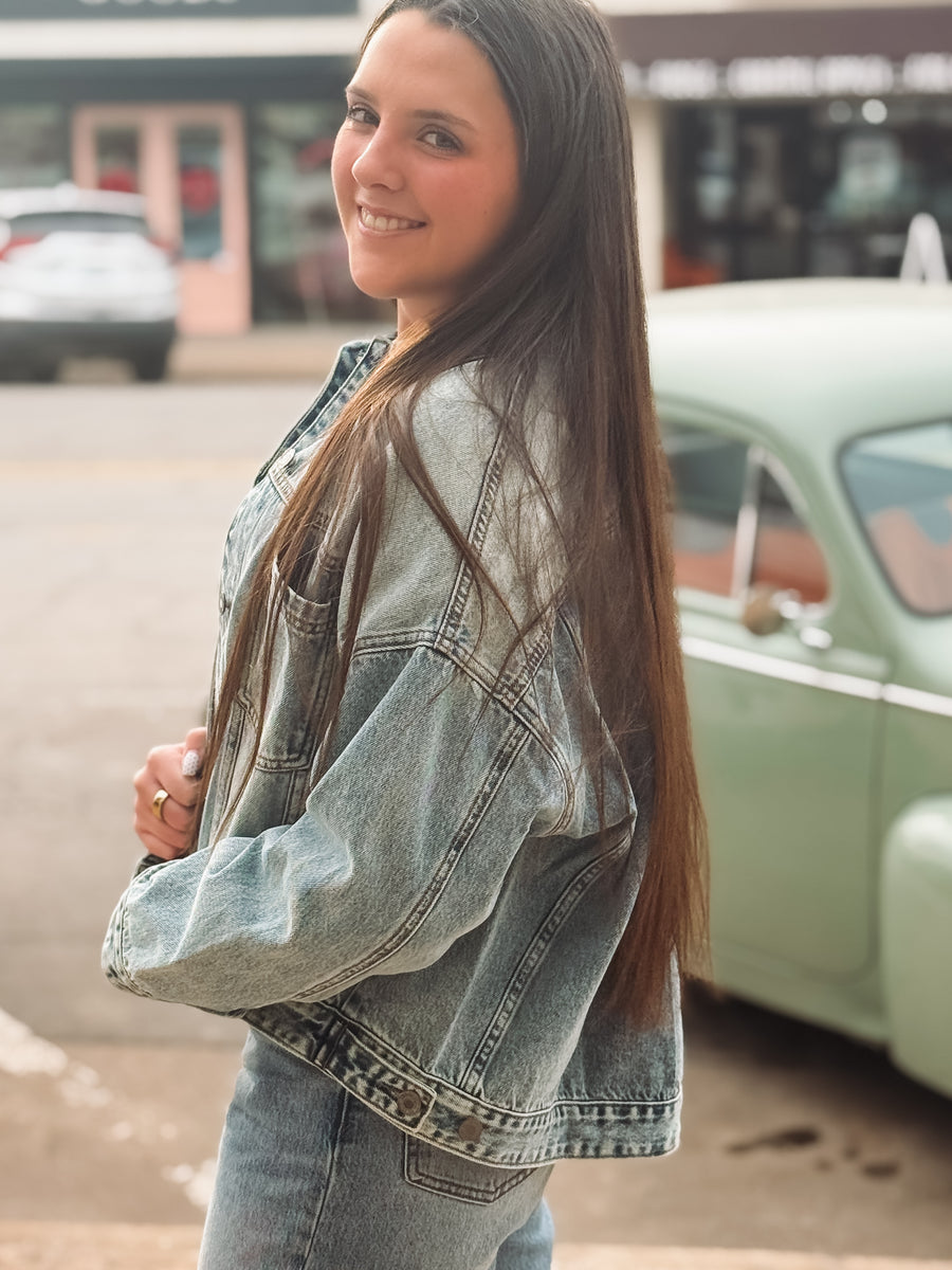Woman wearing a denim jacket standing in front of a vintage car on a street.