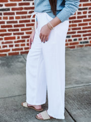 Person wearing light blue sweater and white pants standing against a brick wall.