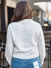 Person wearing a light creamy gray sweater with a brand tag, standing outdoors.