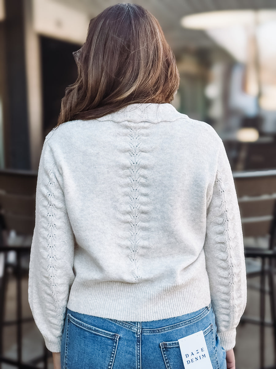 Person wearing a light creamy gray sweater with a brand tag, standing outdoors.