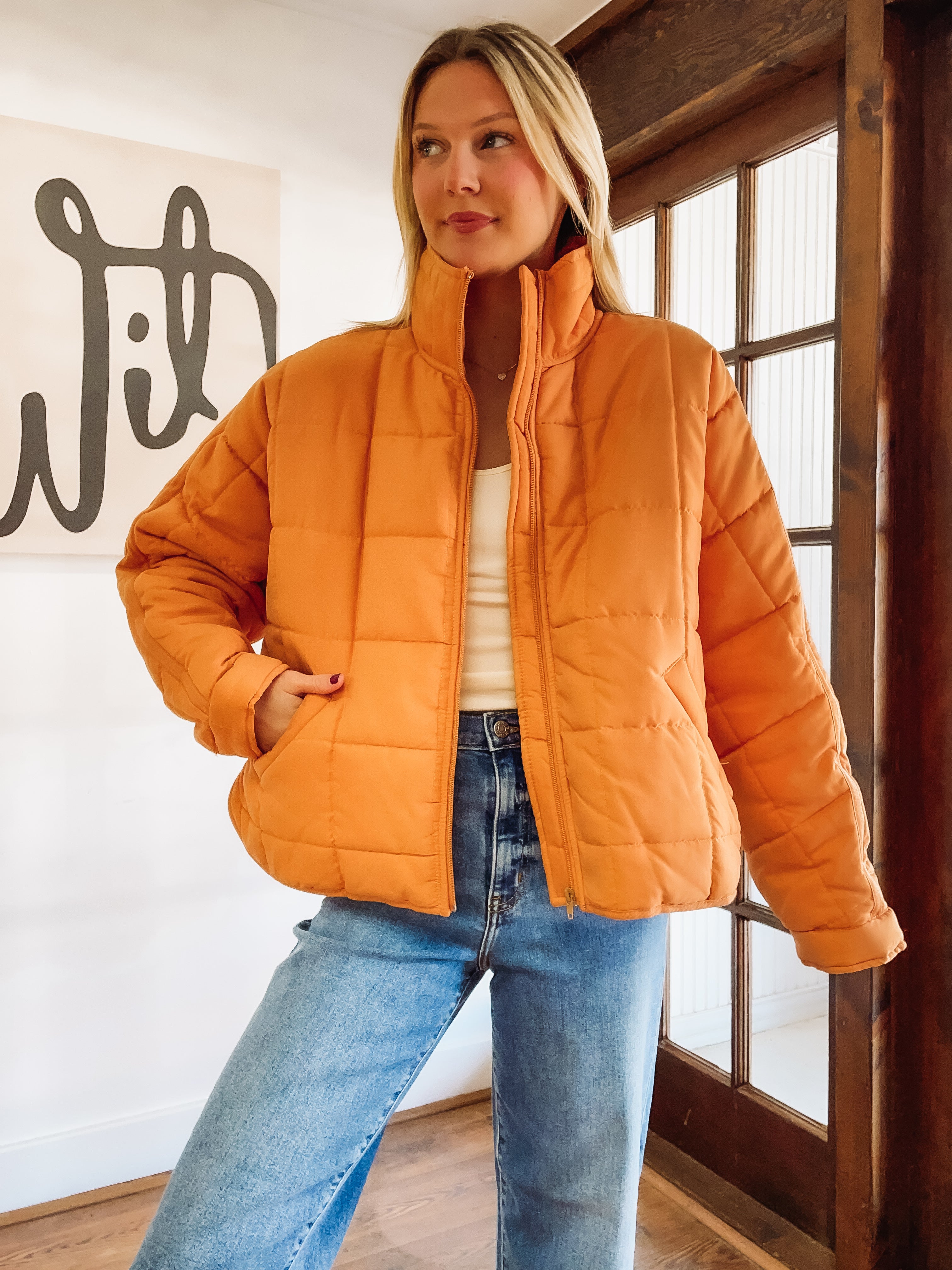 Woman wearing an orange puffer jacket and blue jeans indoors.