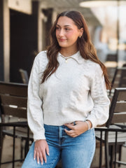 Woman wearing a light gray and creamy sweater and blue jeans standing outdoors.