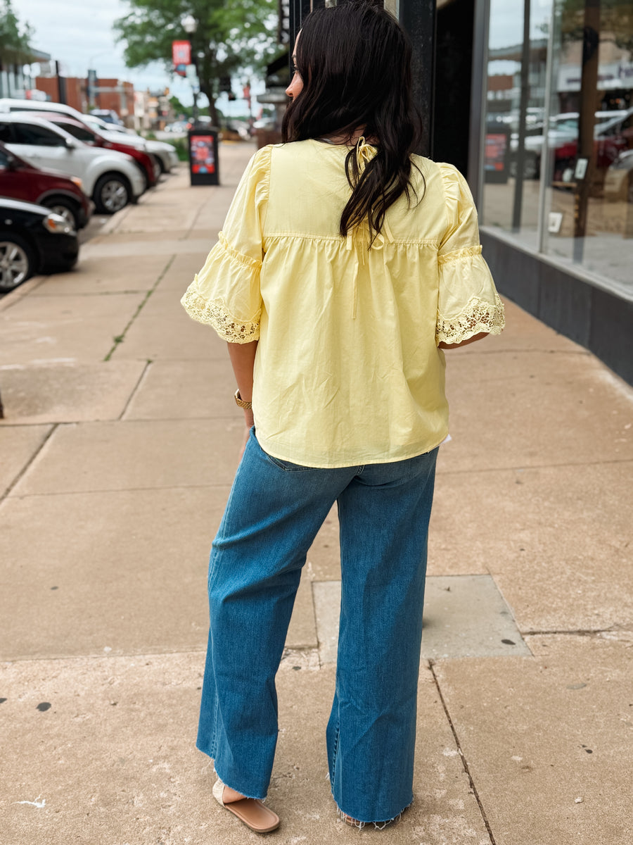 Person wearing a yellow blouse with lace details and blue jeans on a sidewalk.